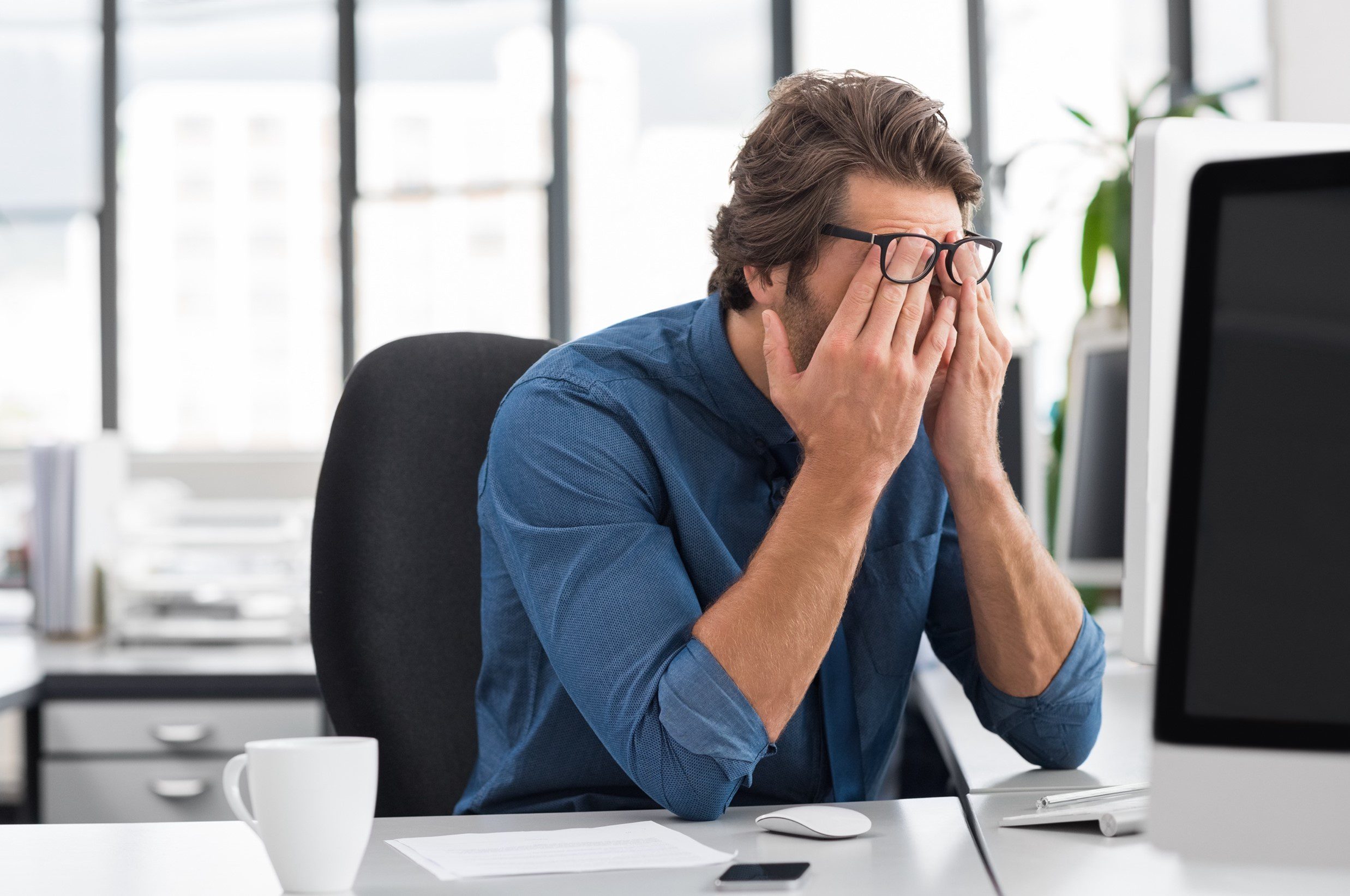 Man with glasses at office desk rubbing his eyes in front of a computer screen